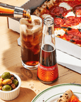 Bottle of Amaro Falso being poured into a glass with a pizza and olives on a wooden table.