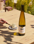 Bottle of wine on a wooden table with a person cutting cheese outdoors.