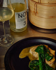 Dinner setting with a dish of food, a glass of white wine, and a bottle labeled 'Noma' on a wooden table.