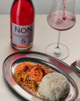 Bottle of 'NON' pink wine with a glass being filled, alongside a dish of food on a table.