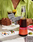 Person holding a glass of dark liquid and playing cards with a bottle of Amaro Falso on a table.