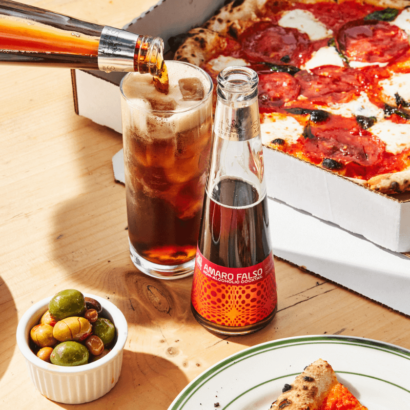 Bottle of Amaro Falso being poured into a glass with a pizza and olives on a wooden table.