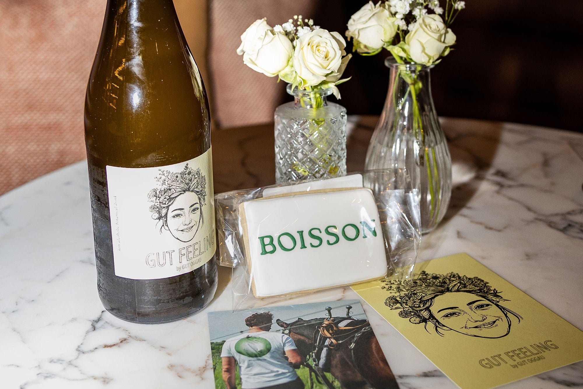 Wine bottle with a label, two small floral arrangements, and cards on a marble table.