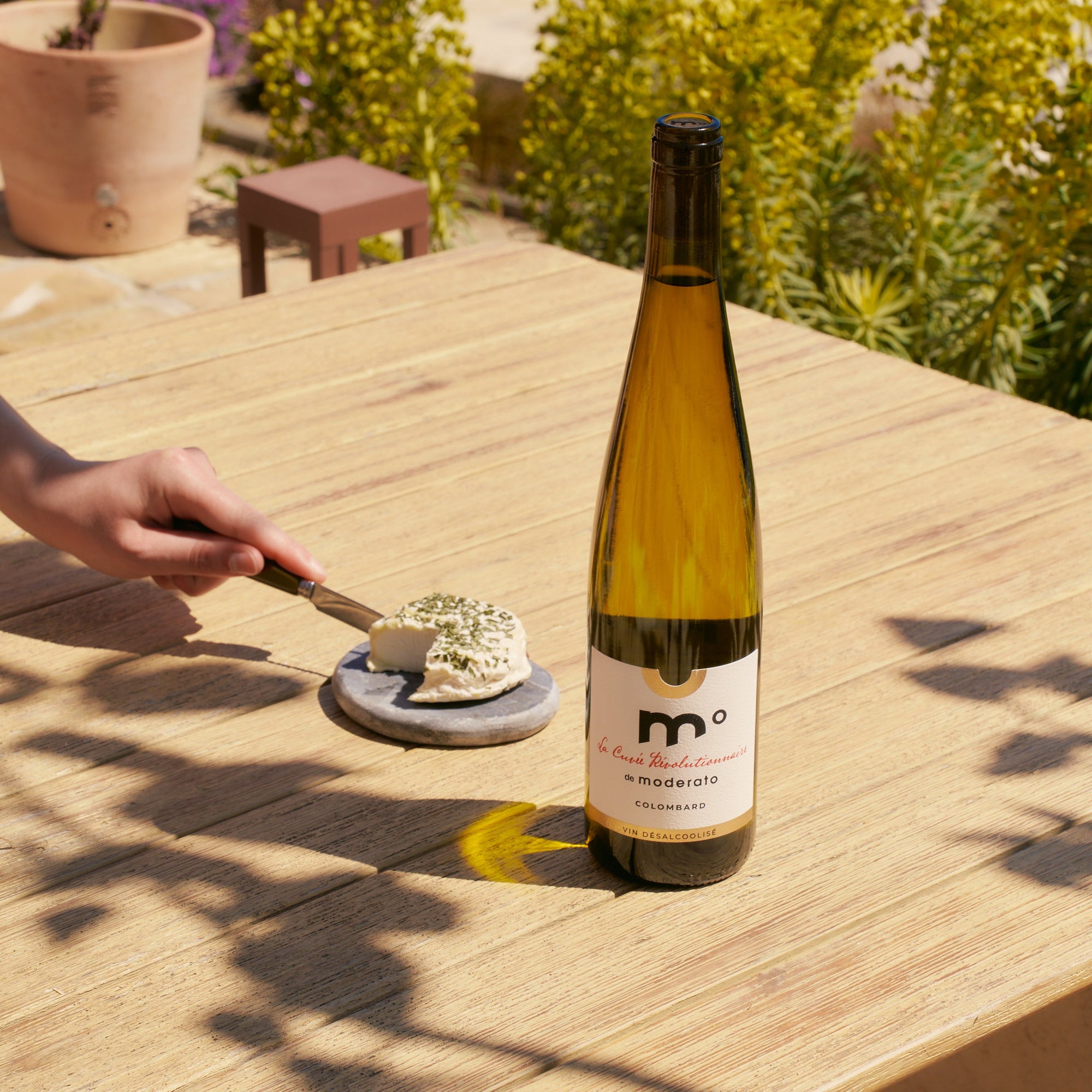 Bottle of wine on a wooden table with a person cutting cheese outdoors.