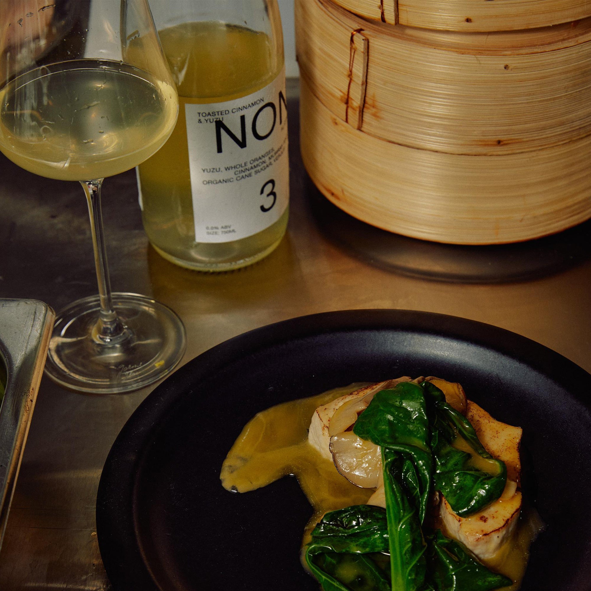 Dinner setting with a dish of food, a glass of white wine, and a bottle labeled 'Noma' on a wooden table.