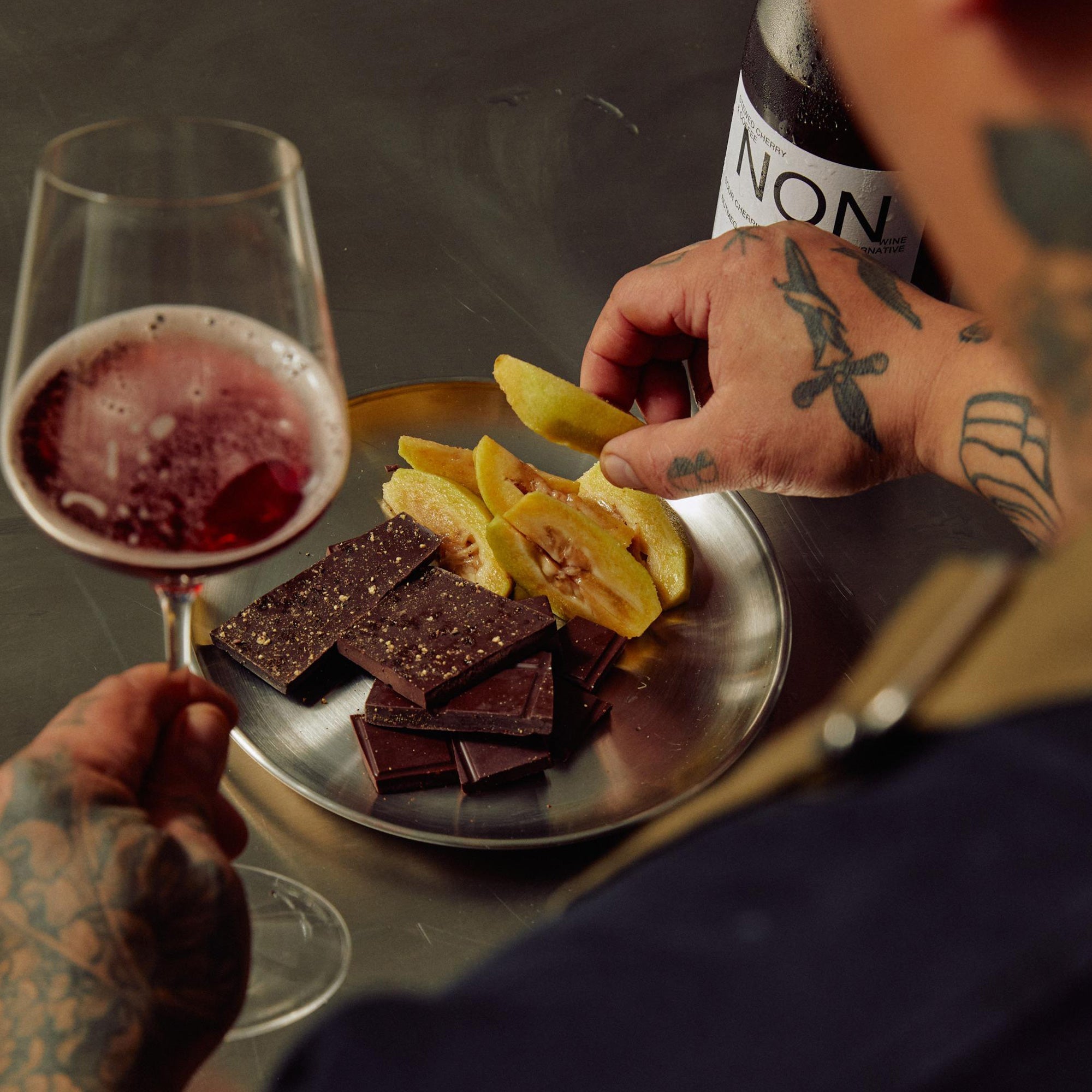 Person holding a glass of red wine with a plate of chocolate bars and fruit, with a bottle labeled 'NON' in the background.