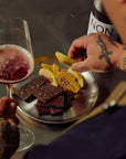 Person holding a glass of red wine with a plate of chocolate bars and fruit, with a bottle labeled 'NON' in the background.