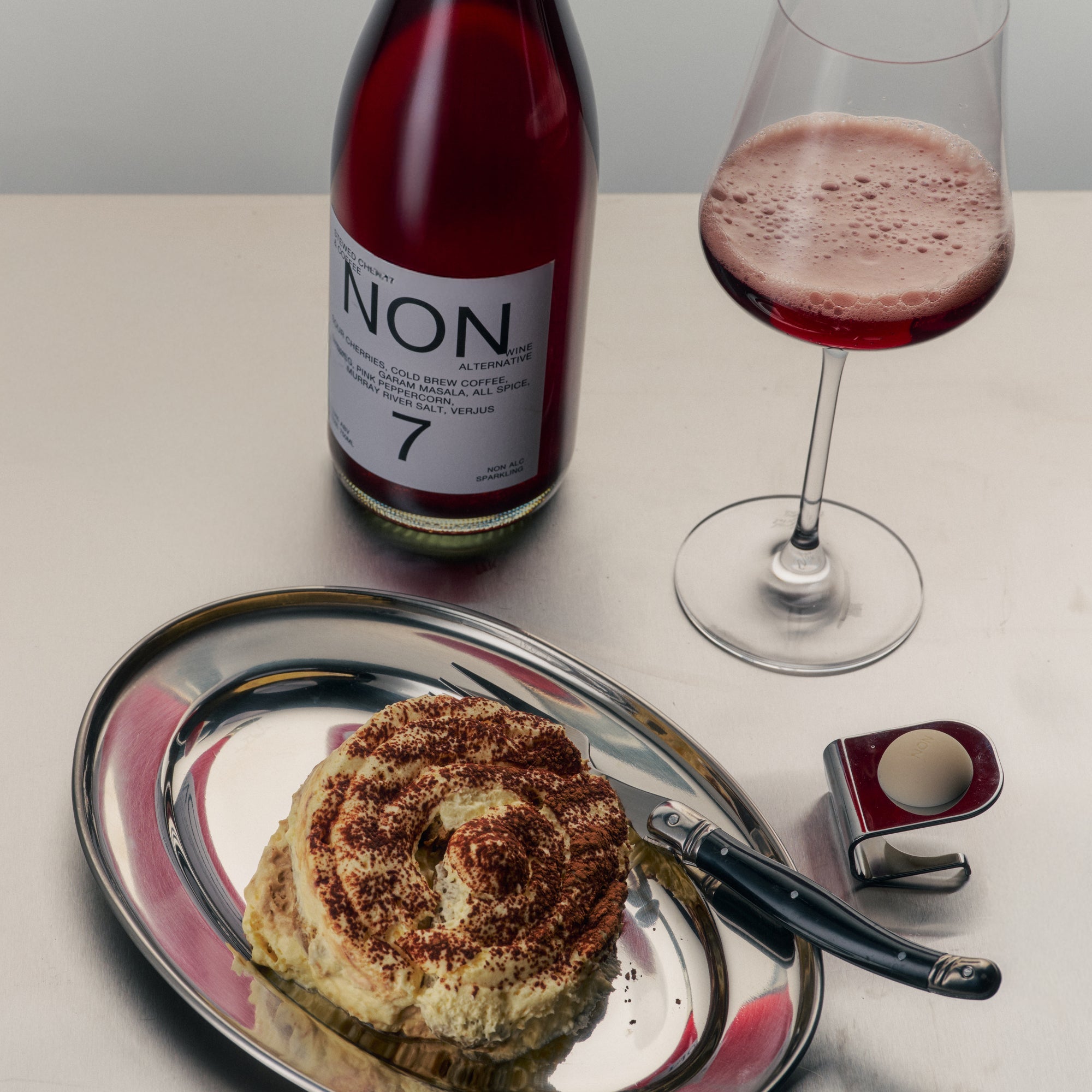 Bottle of NON 7 wine, glass of red wine, and cinnamon roll on a tray.
