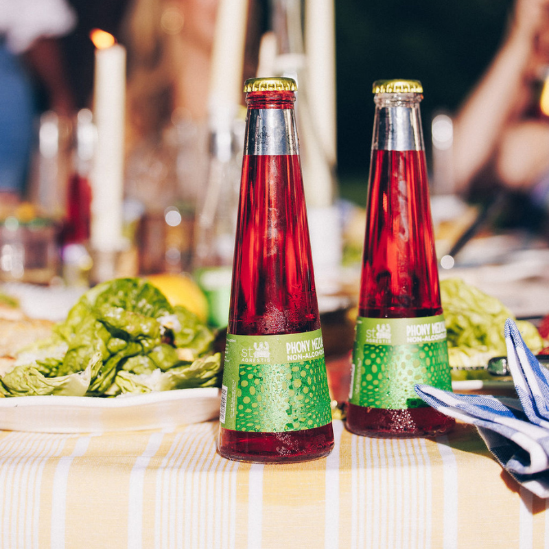 Two bottles of red juice with green labels on a table with food in the background
