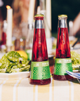 Two bottles of red juice with green labels on a table with food in the background