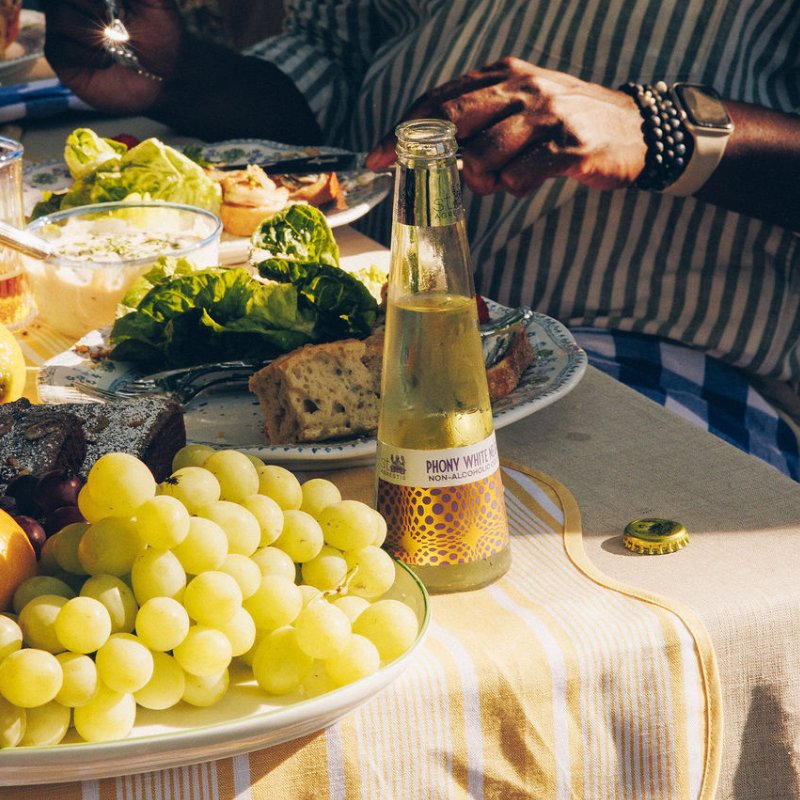 Table with grapes, bread, and a bottle of Phony White wine on a striped tablecloth.