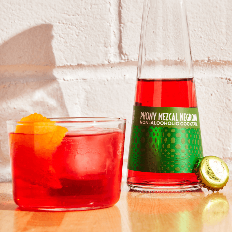Non-alcoholic cocktail in a glass with a bottle of &#39;Phony Mezcal Negroni&#39; on a light brick background