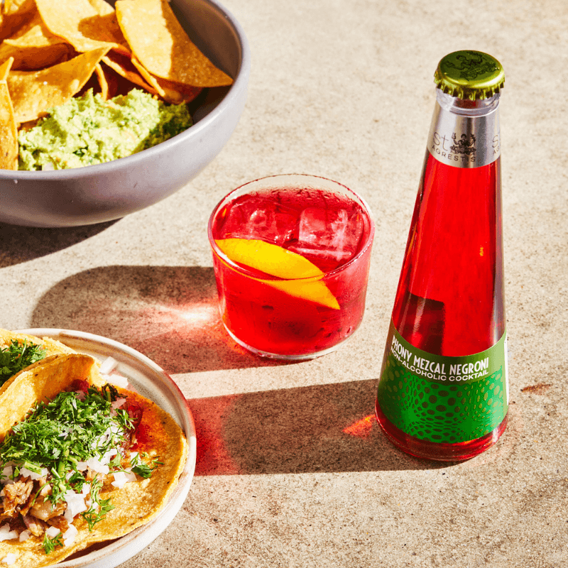 Phony Mezcal Negroni in a glass and a bottle on a concrete surface with Mexican food.