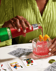 Person pouring Phony Negroni from a bottle into a glass with ice and an orange slice, surrounded by playing cards on a white surface.