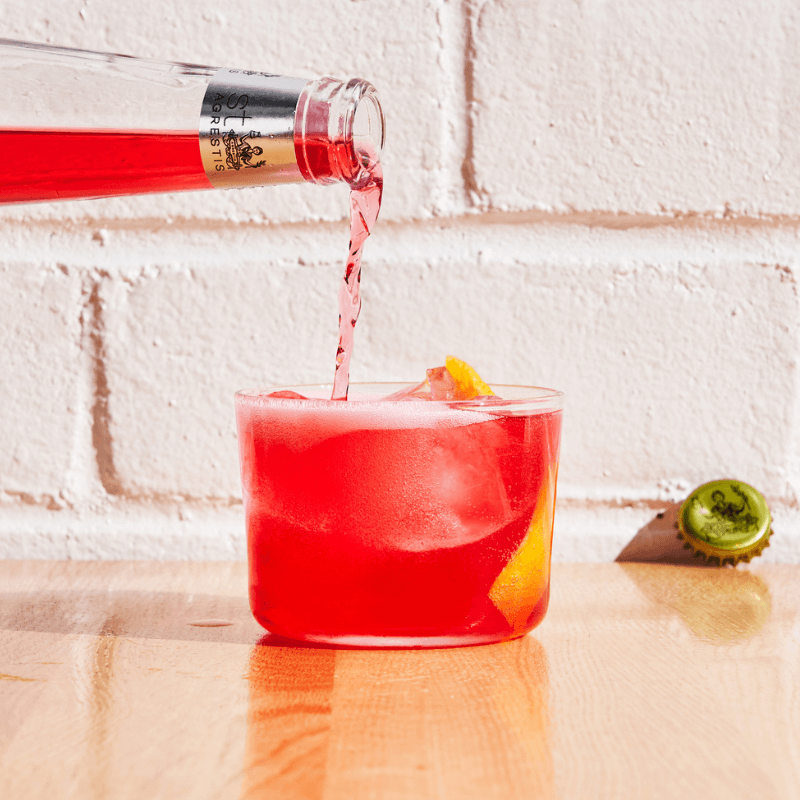 Phony Negroni being poured into a glass with ice and a bottle cap on a wooden surface.