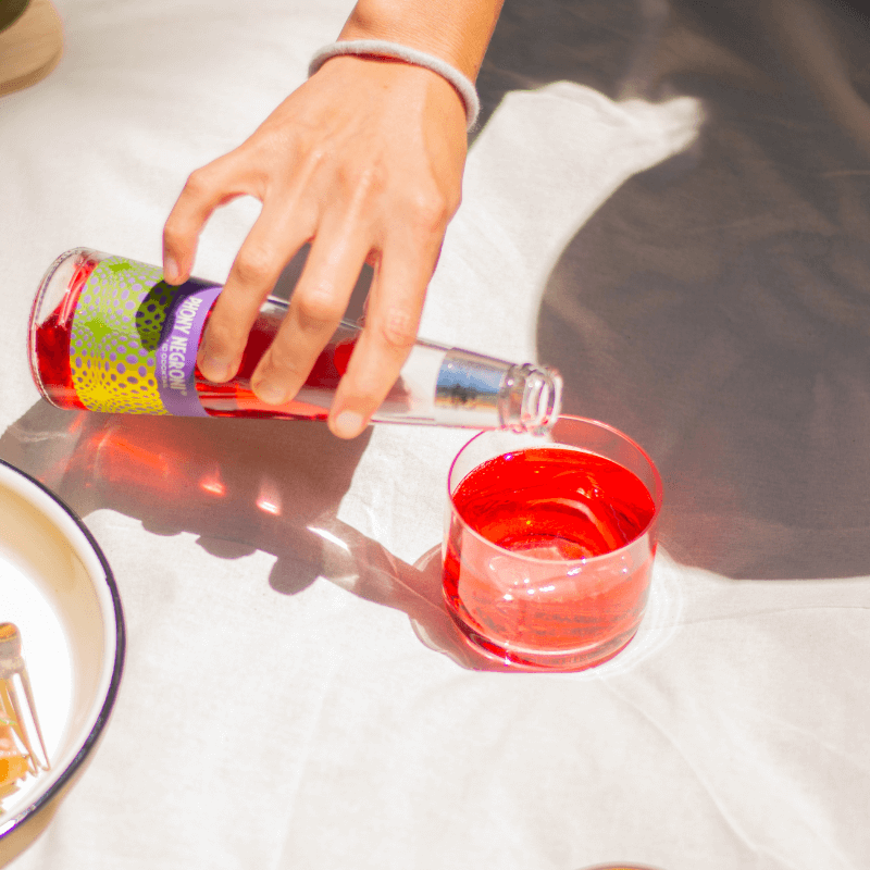 Hand pouring phony negroni from a colorful bottle into a glass on a white surface.