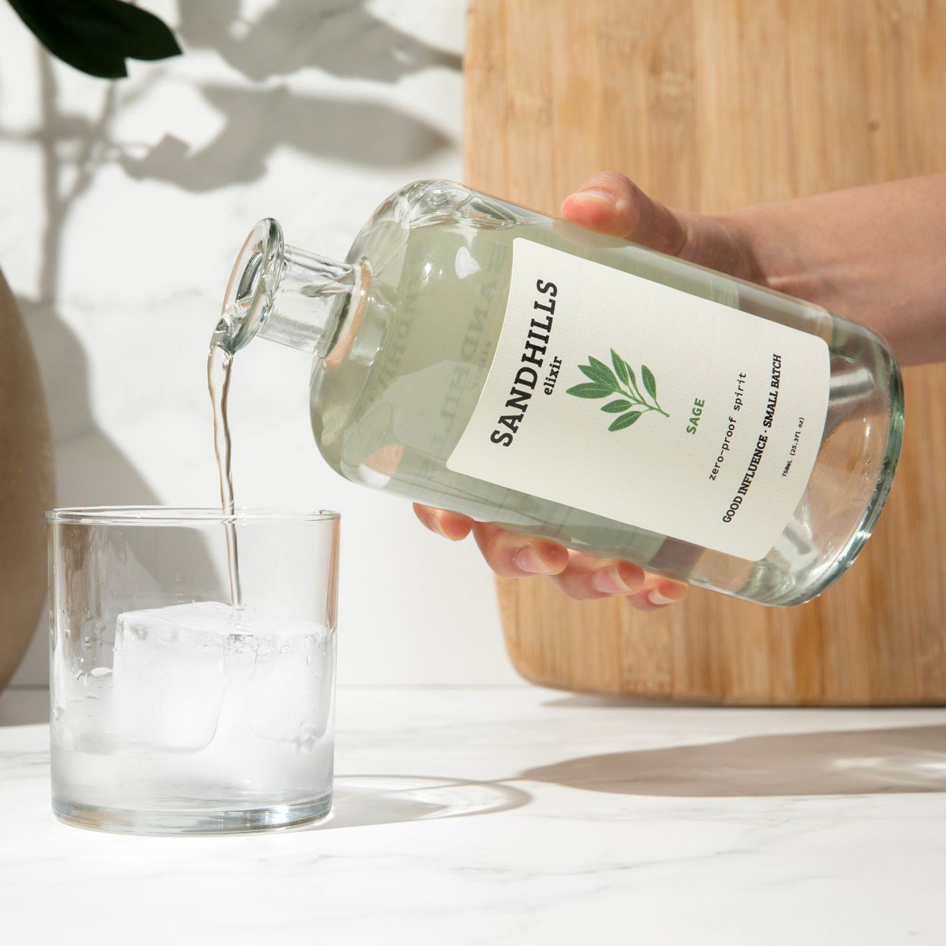 Person pouring liquid from a &#39;Sandhills&#39; bottle into a glass with ice on a light background.