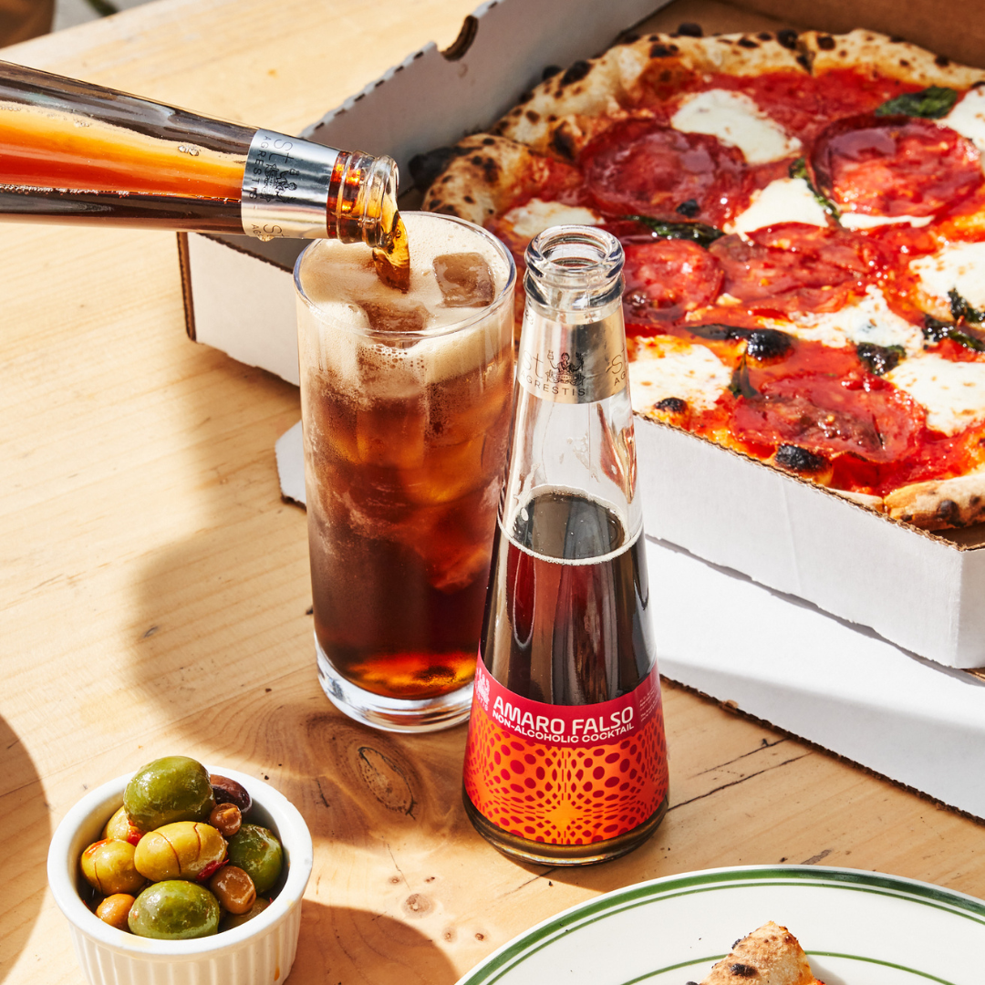 A bottle of Amaro Falso being poured into a glass with a pizza and olives on a wooden table.