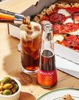 A bottle of Amaro Falso being poured into a glass with a pizza and olives on a wooden table.