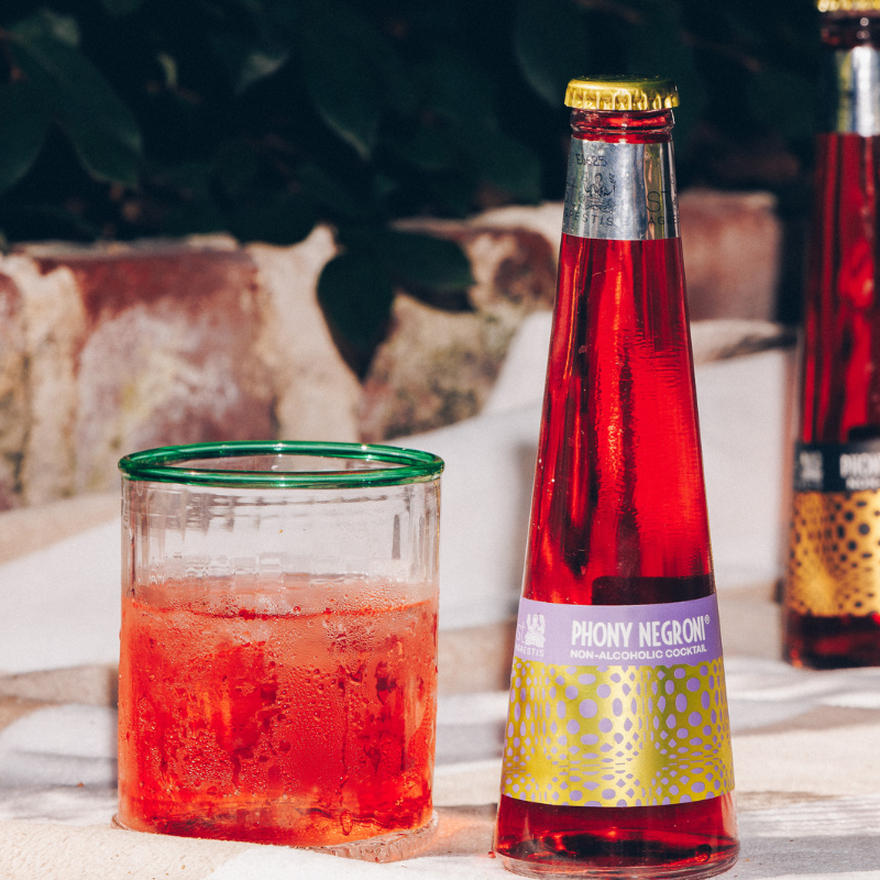 Bottle and glass of red cocktail with &#39;Phony Negroni&#39; label on a blurred outdoor background