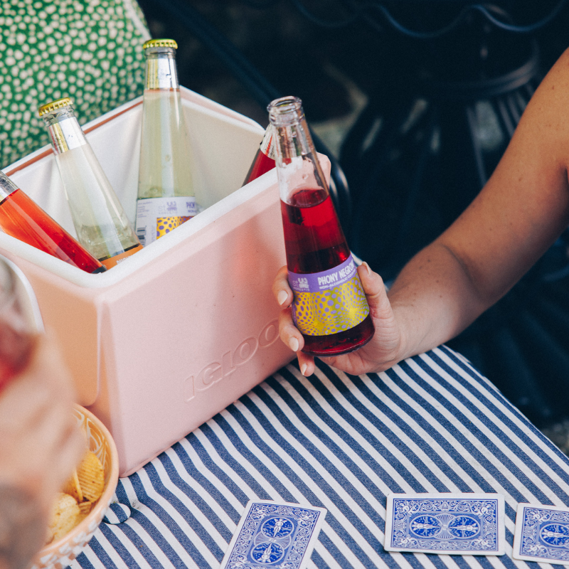 Person holding a bottle of Fentimans at a table with a pink cooler, snacks, and playing cards.