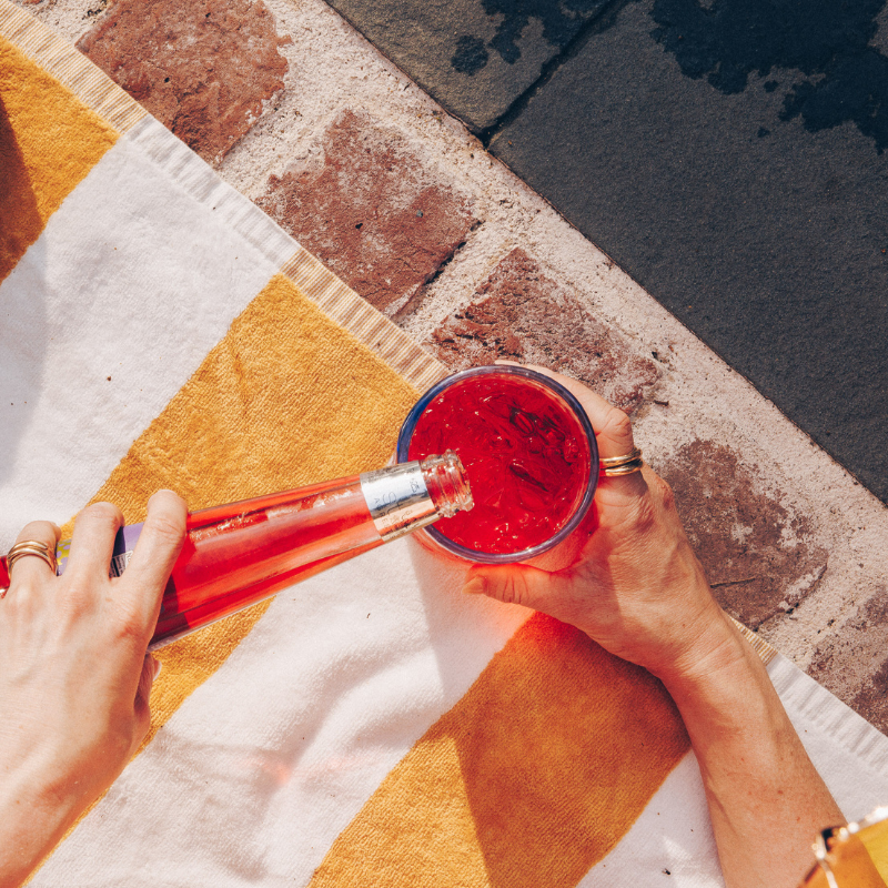 Person pouring red liquid from a bottle into a glass on a colorful outdoor mat.