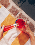 Person pouring red liquid from a bottle into a glass on a colorful outdoor mat.
